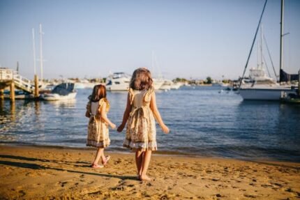 Twin sisters in matching dresses playing on a sunny beach near moored yachts.