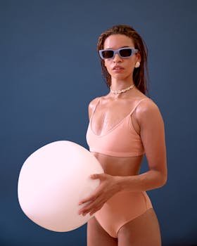 Model in stylish swimwear with sunglasses and inflatable pillow in studio setting.